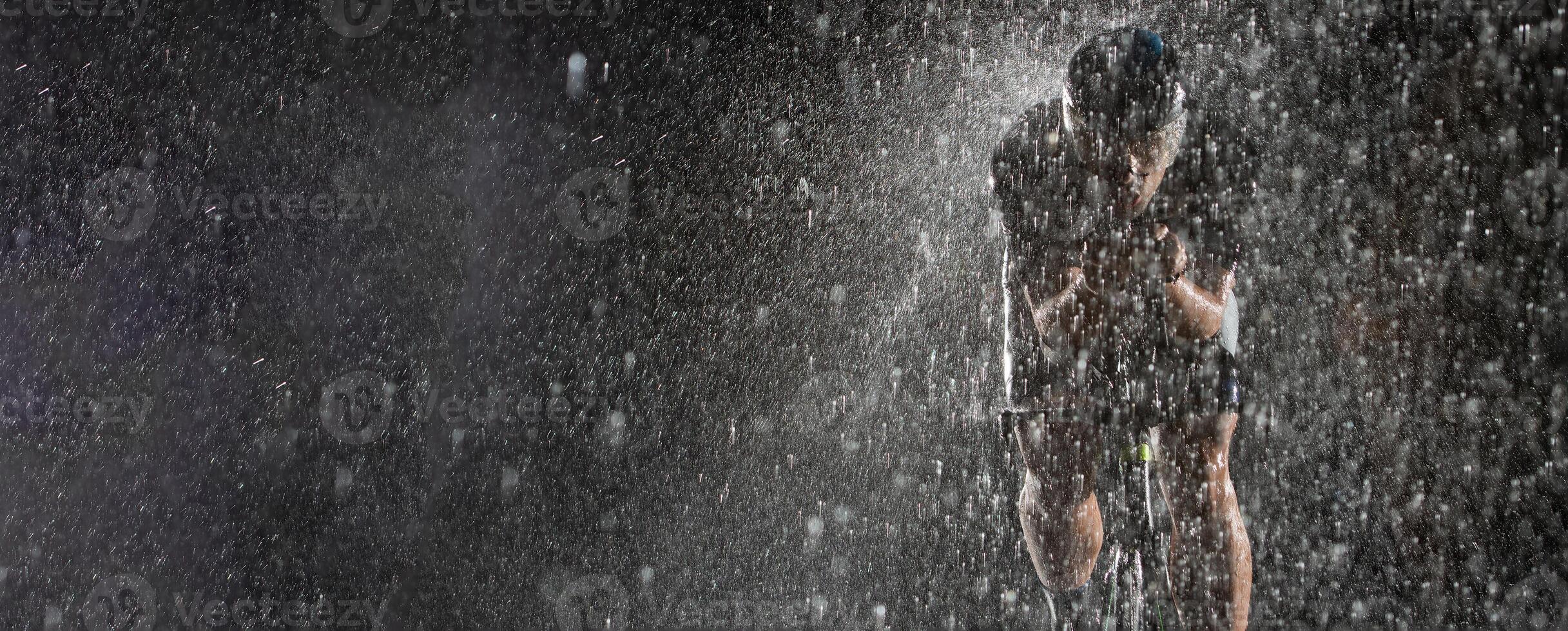 A triathlete braving the rain as he cycles through the night, preparing himself for the upcoming marathon. The blurred raindrops in the foreground and the dark, moody atmosphere in the background add photo