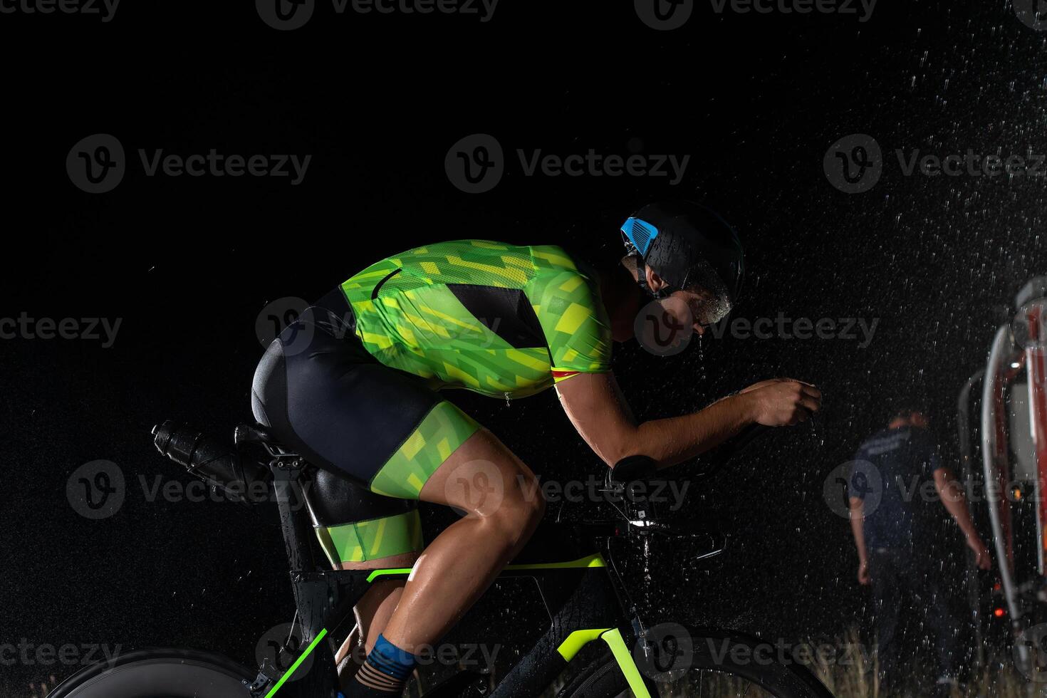 A triathlete braving the rain as he cycles through the night, preparing himself for the upcoming marathon. The blurred raindrops in the foreground and the dark, moody atmosphere in the background add photo
