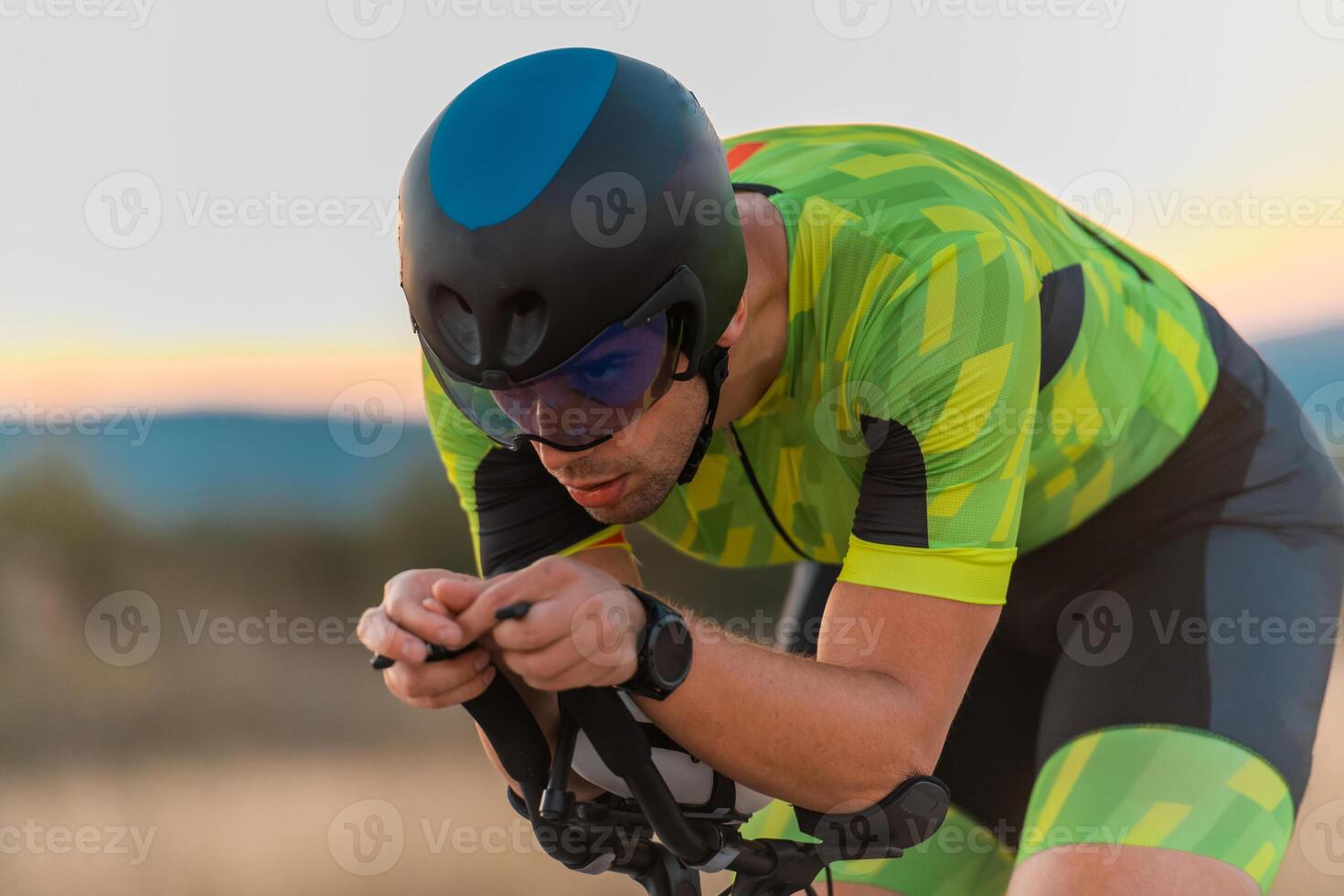 Close up photo of triathlete riding his bicycle during sunset, preparing for a marathon. The warm colors of the sky provide a beautiful backdrop for his determined and focused effort.