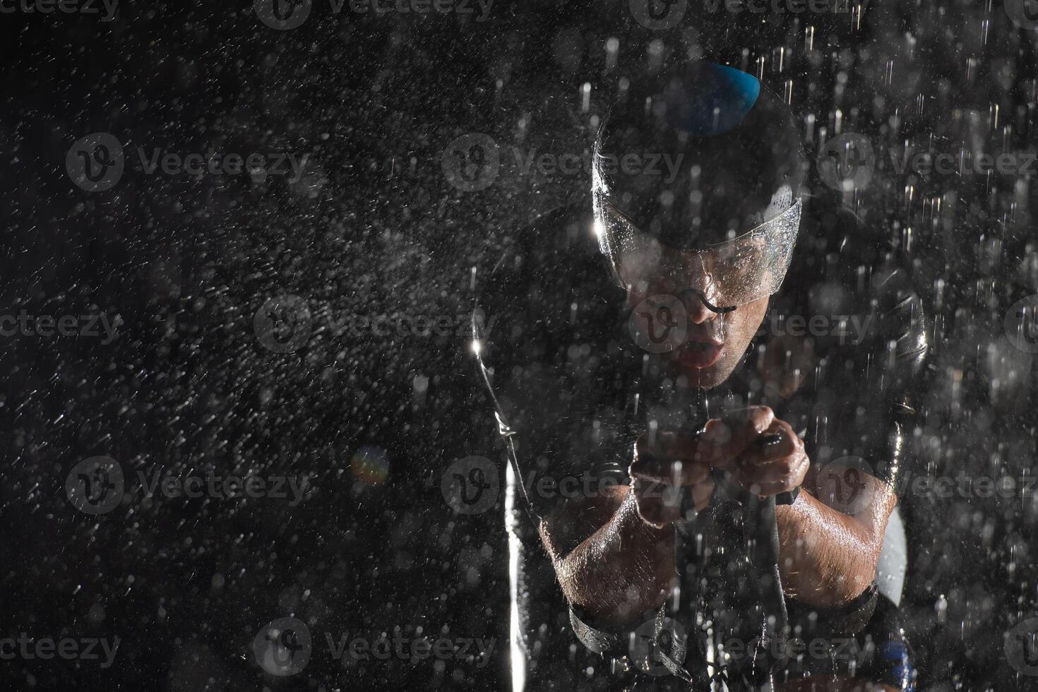 A triathlete braving the rain as he cycles through the night, preparing himself for the upcoming marathon. The blurred raindrops in the foreground and the dark, moody atmosphere in the background add photo