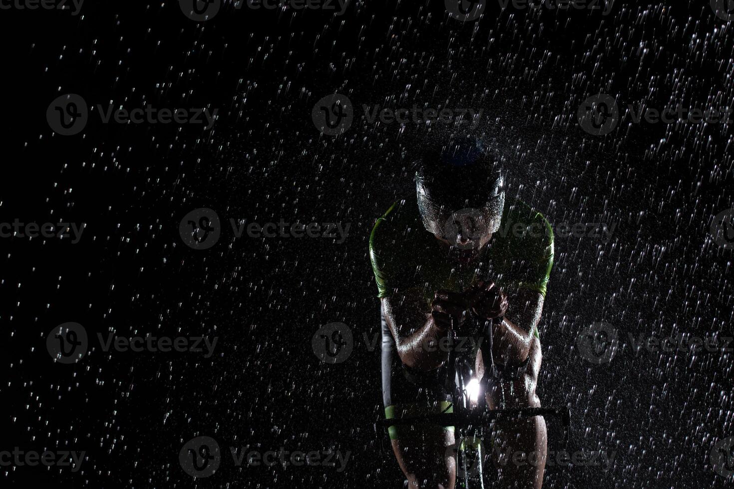 A triathlete braving the rain as he cycles through the night, preparing himself for the upcoming marathon. The blurred raindrops in the foreground and the dark, moody atmosphere in the background add photo
