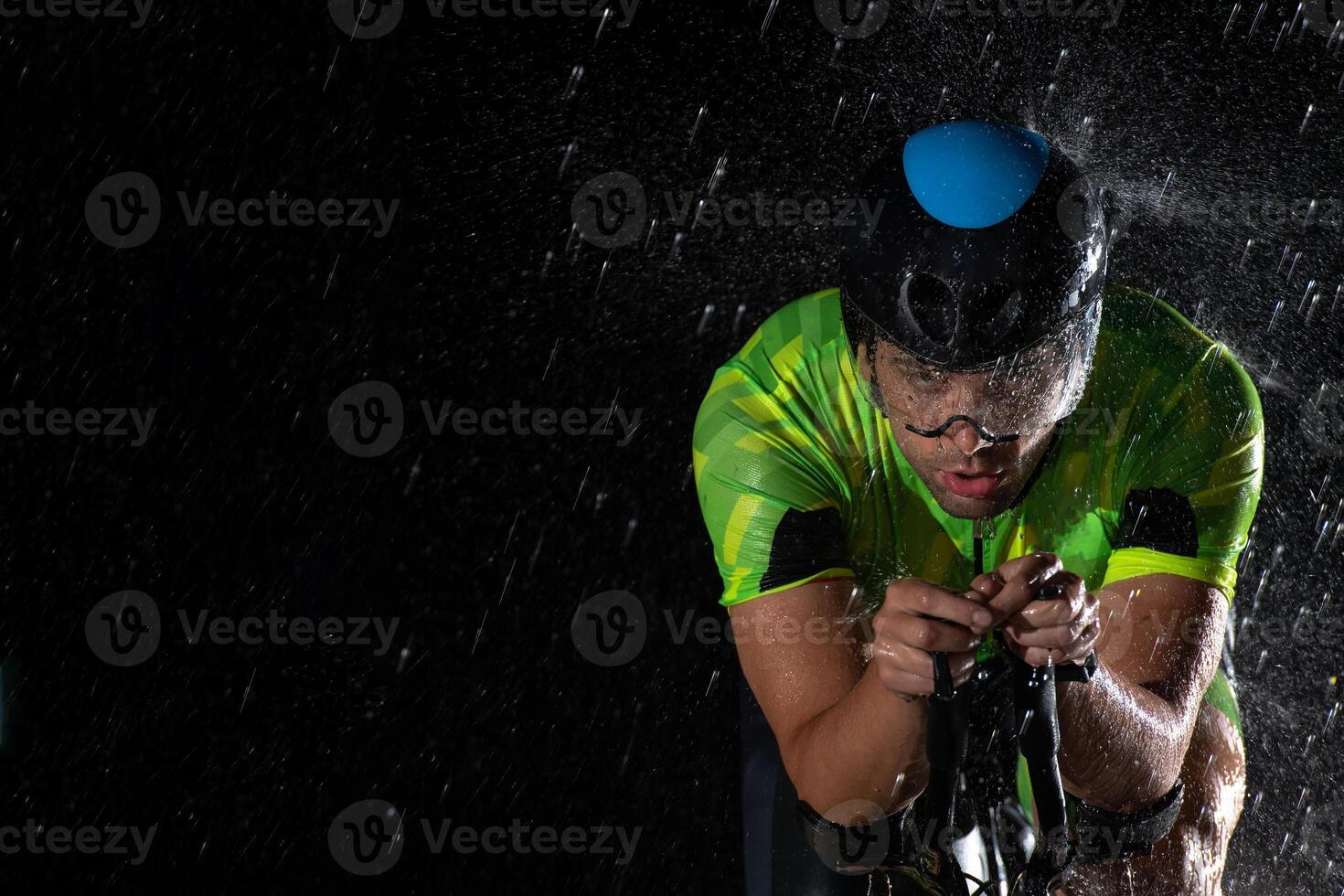 A triathlete braving the rain as he cycles through the night, preparing himself for the upcoming marathon. The blurred raindrops in the foreground and the dark, moody atmosphere in the background add photo