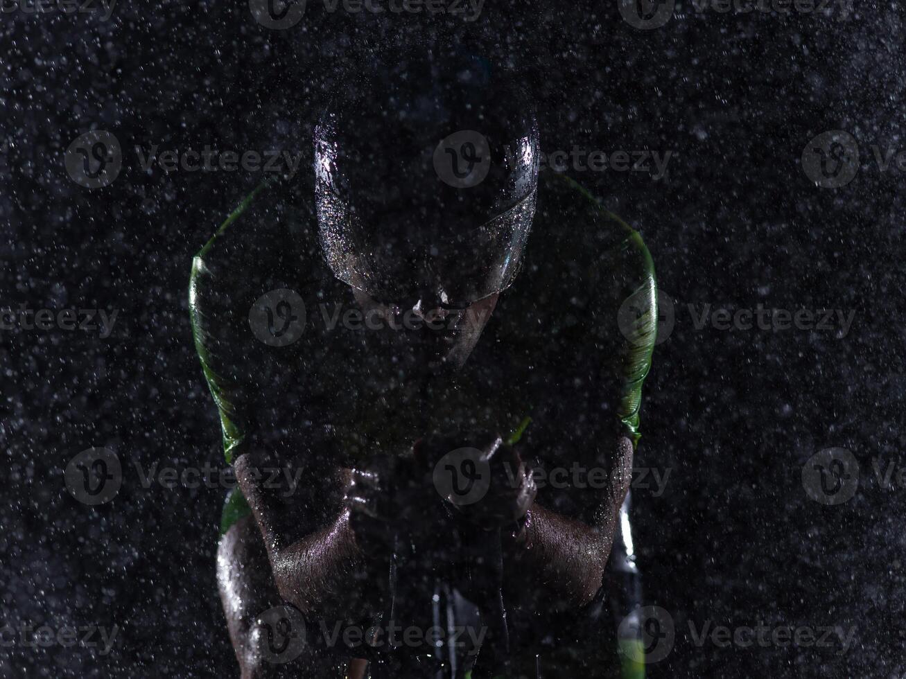 A triathlete braving the rain as he cycles through the night, preparing himself for the upcoming marathon. The blurred raindrops in the foreground and the dark, moody atmosphere in the background add photo