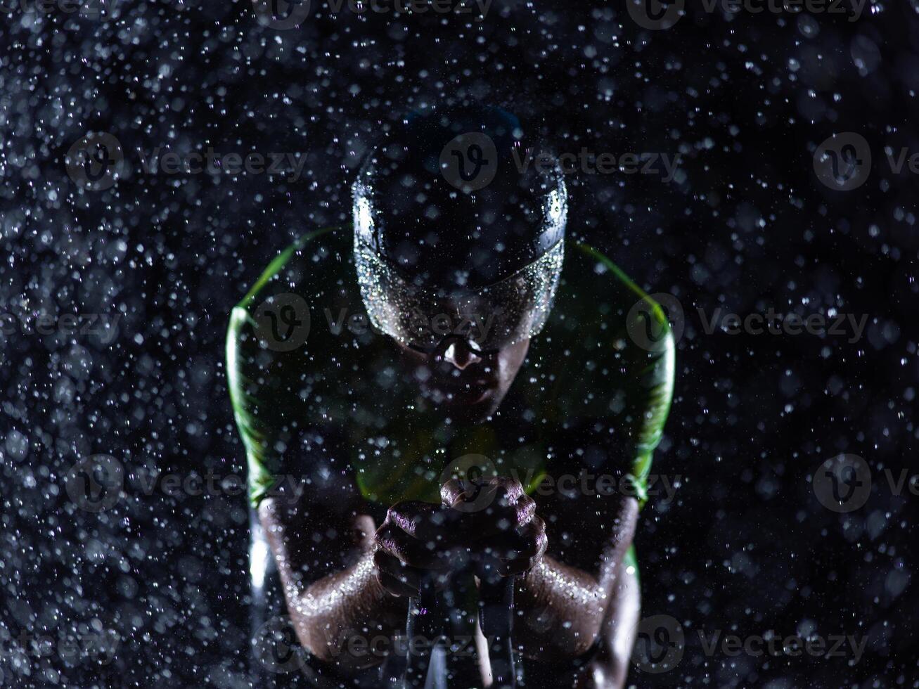 A triathlete braving the rain as he cycles through the night, preparing himself for the upcoming marathon. The blurred raindrops in the foreground and the dark, moody atmosphere in the background add photo
