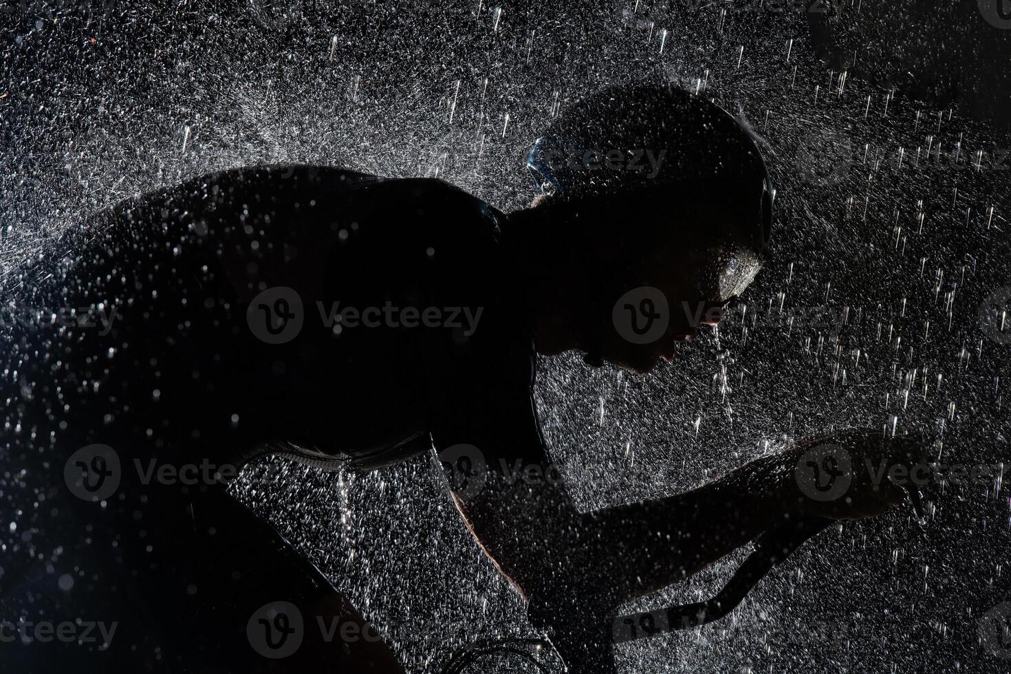 A triathlete braving the rain as he cycles through the night, preparing himself for the upcoming marathon. The blurred raindrops in the foreground and the dark, moody atmosphere in the background add photo