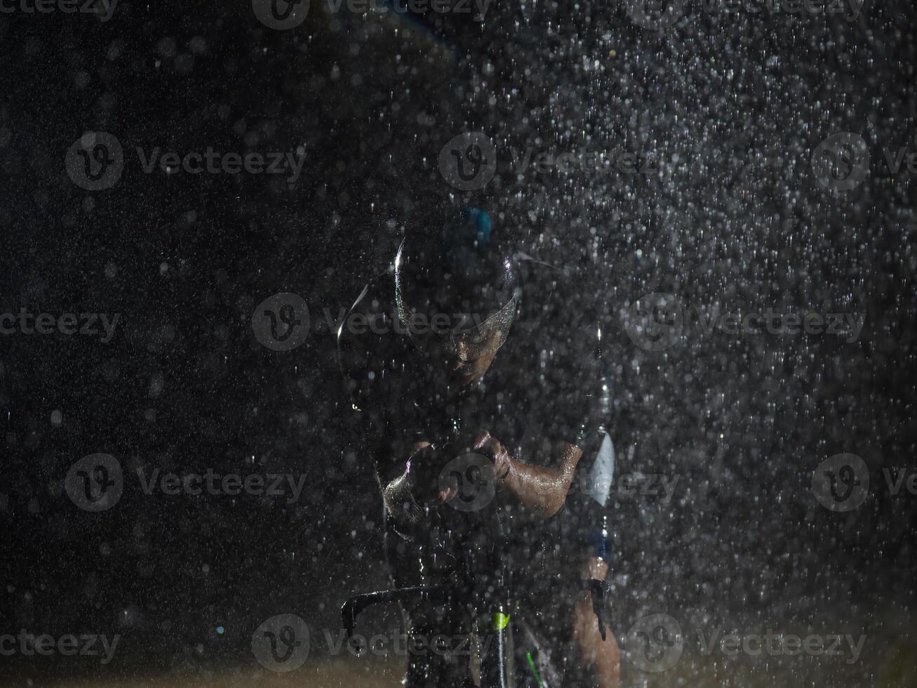 A triathlete braving the rain as he cycles through the night, preparing himself for the upcoming marathon. The blurred raindrops in the foreground and the dark, moody atmosphere in the background add photo