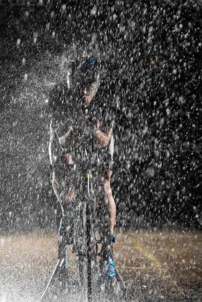 A triathlete braving the rain as he cycles through the night, preparing himself for the upcoming marathon. The blurred raindrops in the foreground and the dark, moody atmosphere in the background add photo