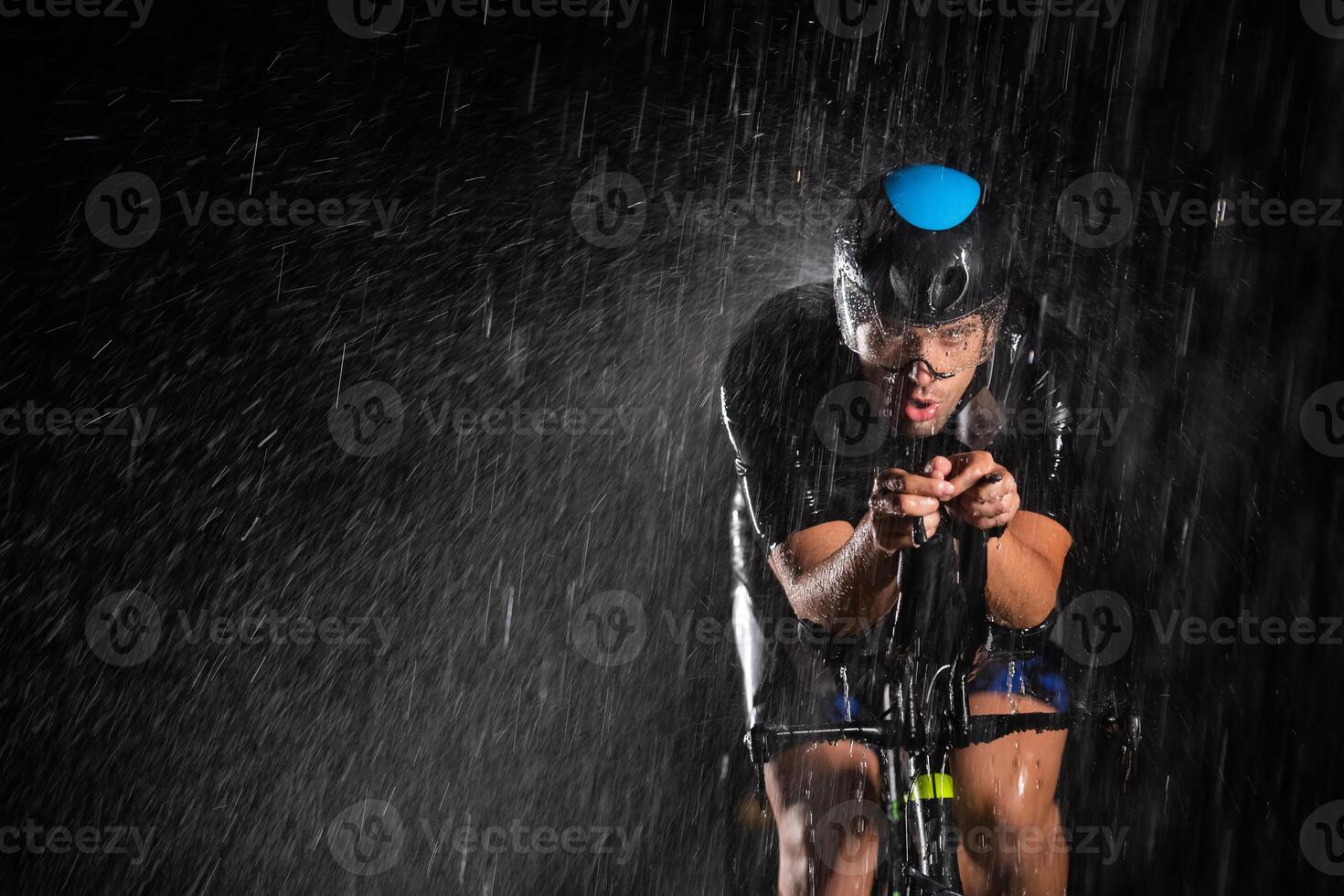 A triathlete braving the rain as he cycles through the night, preparing himself for the upcoming marathon. The blurred raindrops in the foreground and the dark, moody atmosphere in the background add photo