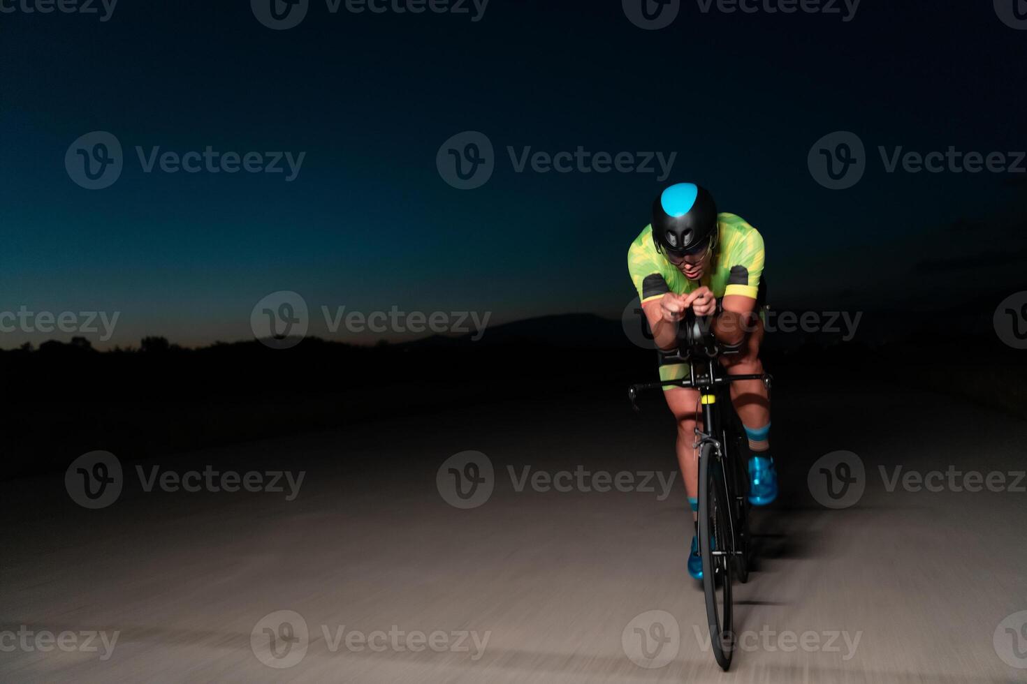A triathlete rides his bike in the darkness of night, pushing himself to prepare for a marathon. The contrast between the darkness and the light of his bike creates a sense of drama and highlights the photo