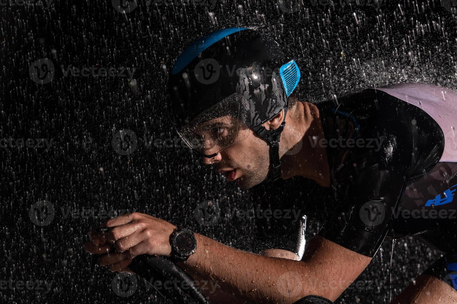 A triathlete braving the rain as he cycles through the night, preparing himself for the upcoming marathon. The blurred raindrops in the foreground and the dark, moody atmosphere in the background add photo