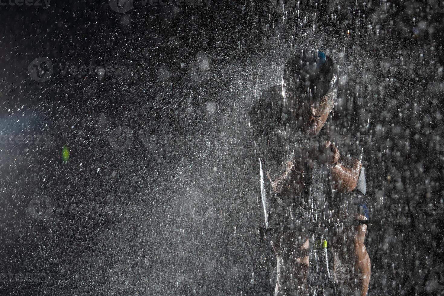 A triathlete braving the rain as he cycles through the night, preparing himself for the upcoming marathon. The blurred raindrops in the foreground and the dark, moody atmosphere in the background add photo