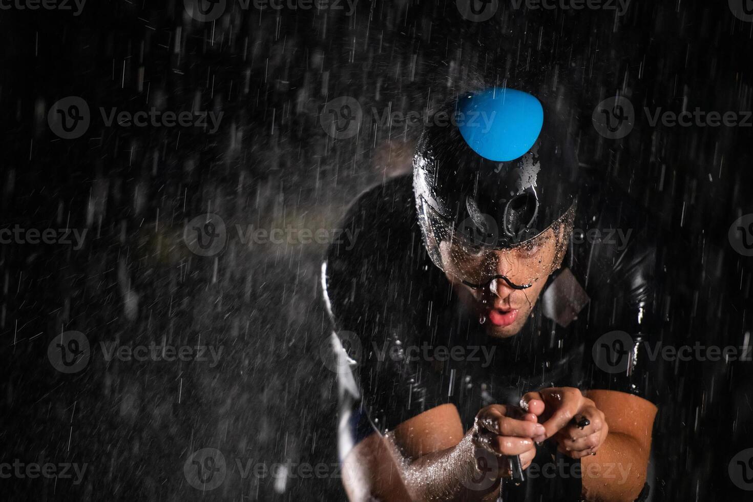 A triathlete braving the rain as he cycles through the night, preparing himself for the upcoming marathon. The blurred raindrops in the foreground and the dark, moody atmosphere in the background add photo