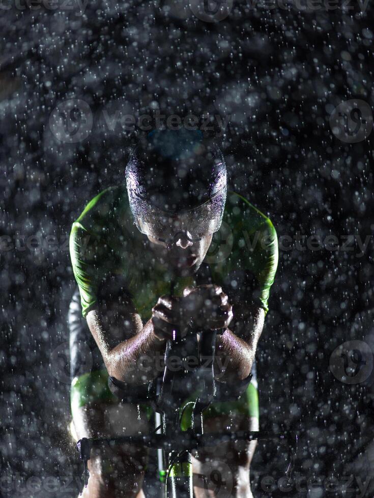A triathlete braving the rain as he cycles through the night, preparing himself for the upcoming marathon. The blurred raindrops in the foreground and the dark, moody atmosphere in the background add photo