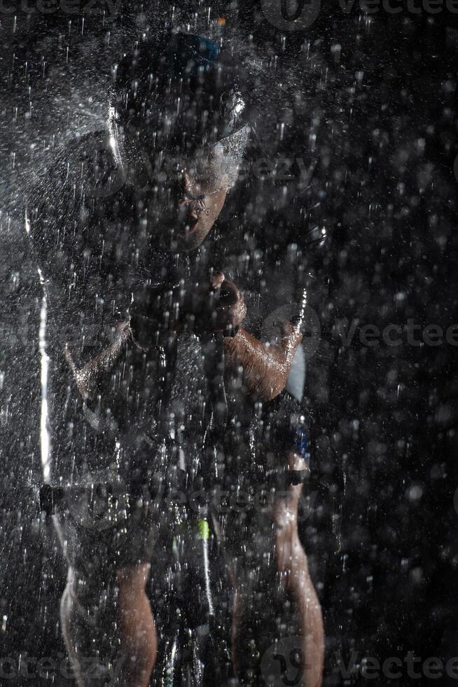 A triathlete braving the rain as he cycles through the night, preparing himself for the upcoming marathon. The blurred raindrops in the foreground and the dark, moody atmosphere in the background add photo