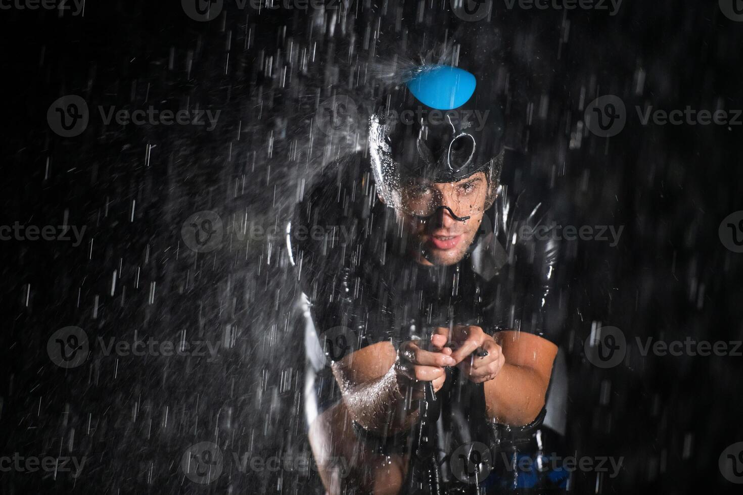 A triathlete braving the rain as he cycles through the night, preparing himself for the upcoming marathon. The blurred raindrops in the foreground and the dark, moody atmosphere in the background add photo