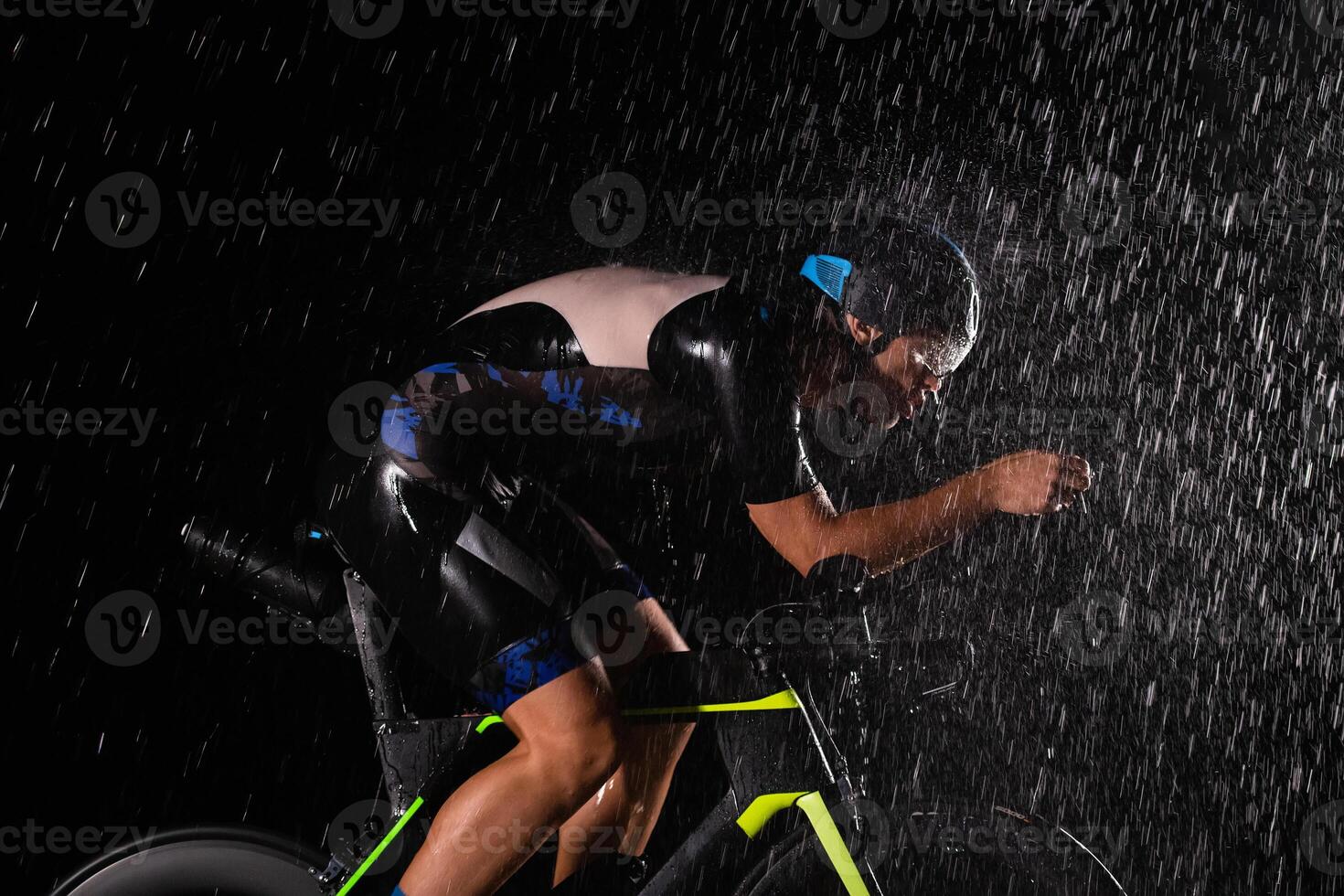 A triathlete braving the rain as he cycles through the night, preparing himself for the upcoming marathon. The blurred raindrops in the foreground and the dark, moody atmosphere in the background add photo