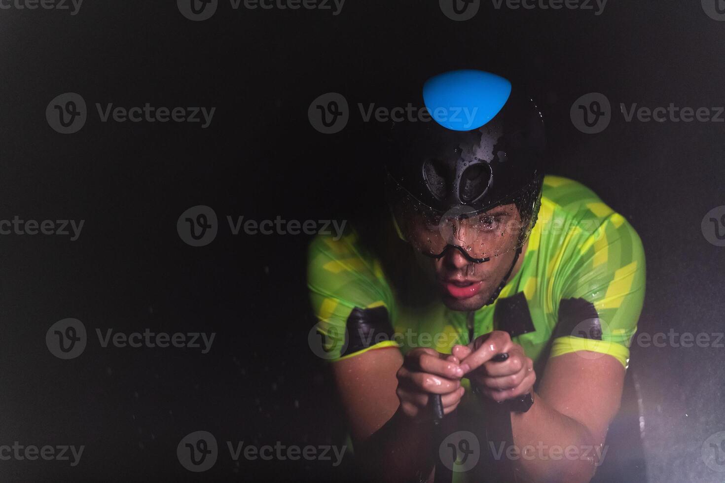A triathlete braving the rain as he cycles through the night, preparing himself for the upcoming marathon. The blurred raindrops in the foreground and the dark, moody atmosphere in the background add photo