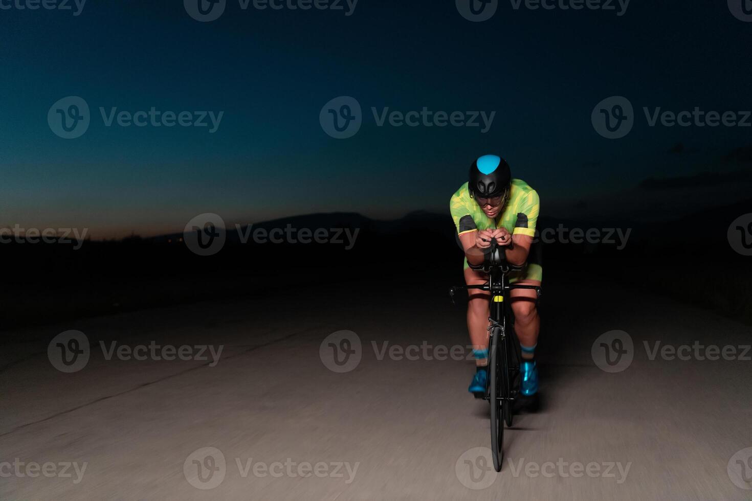 A triathlete rides his bike in the darkness of night, pushing himself to prepare for a marathon. The contrast between the darkness and the light of his bike creates a sense of drama and highlights the photo