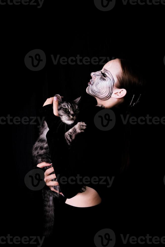 image for Halloween, girl with spiral makeup on her face with a gray cat in her arms. Celebration and pets concept photo