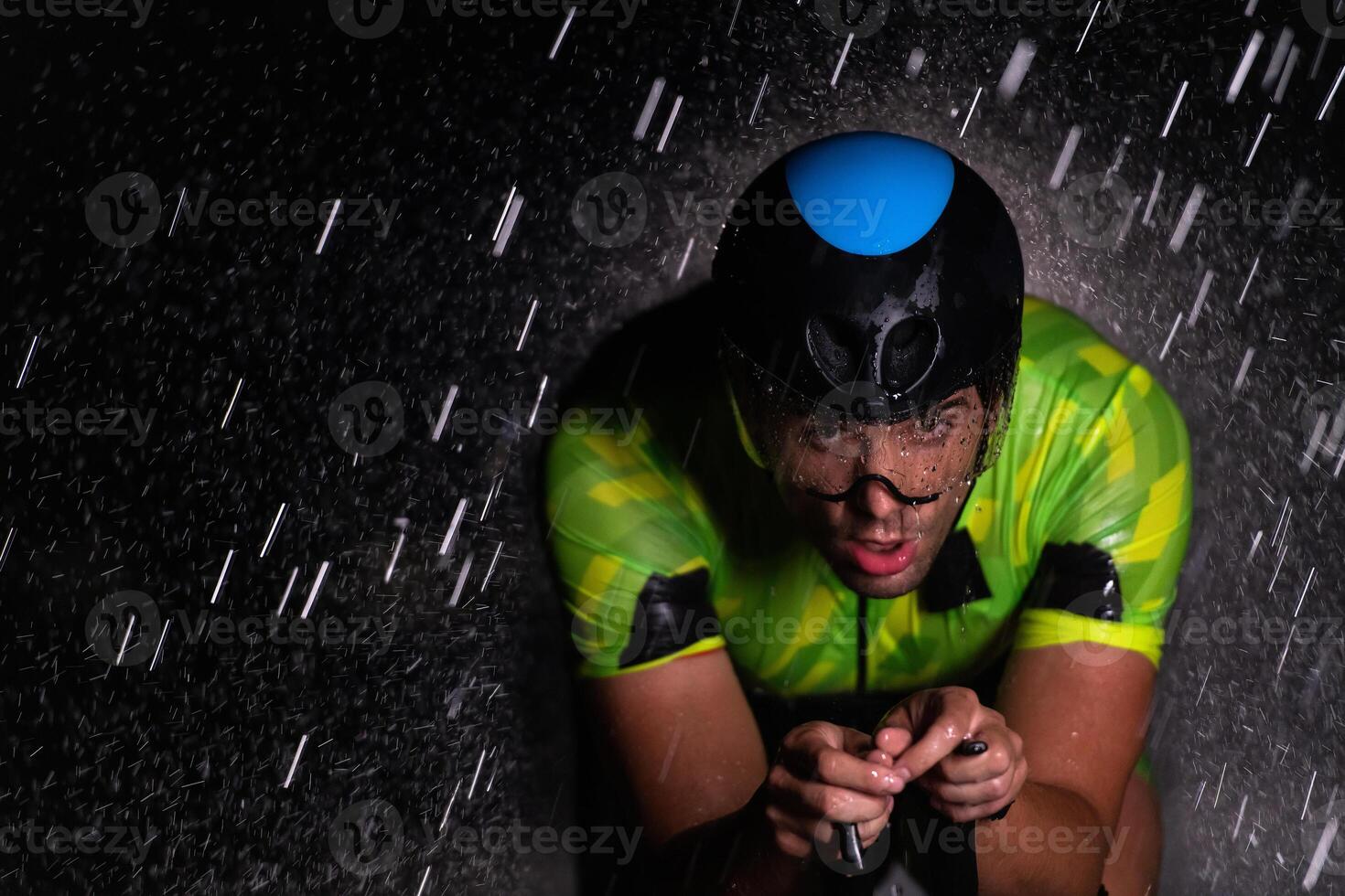 A triathlete braving the rain as he cycles through the night, preparing himself for the upcoming marathon. The blurred raindrops in the foreground and the dark, moody atmosphere in the background add photo