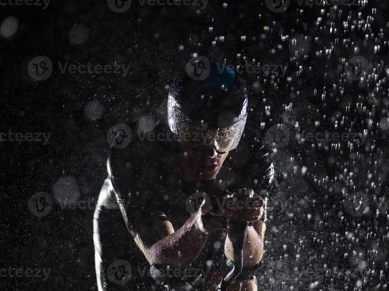 A triathlete braving the rain as he cycles through the night, preparing himself for the upcoming marathon. The blurred raindrops in the foreground and the dark, moody atmosphere in the background add photo