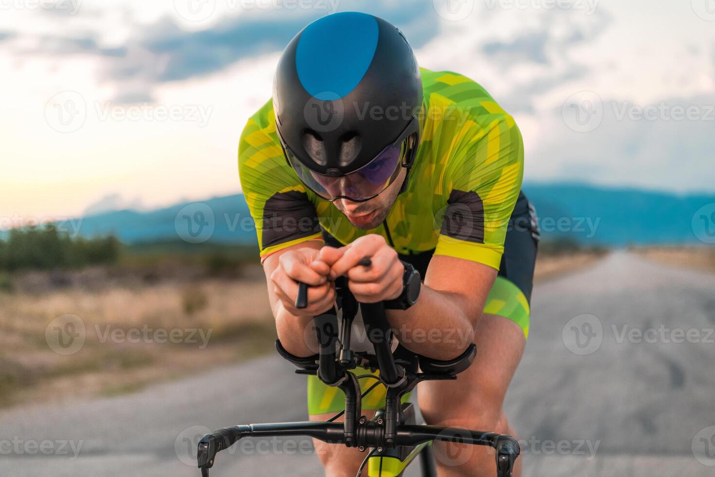 Close up photo of triathlete riding his bicycle during sunset, preparing for a marathon. The warm colors of the sky provide a beautiful backdrop for his determined and focused effort.