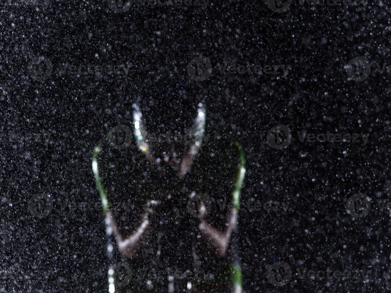 A triathlete braving the rain as he cycles through the night, preparing himself for the upcoming marathon. The blurred raindrops in the foreground and the dark, moody atmosphere in the background add photo