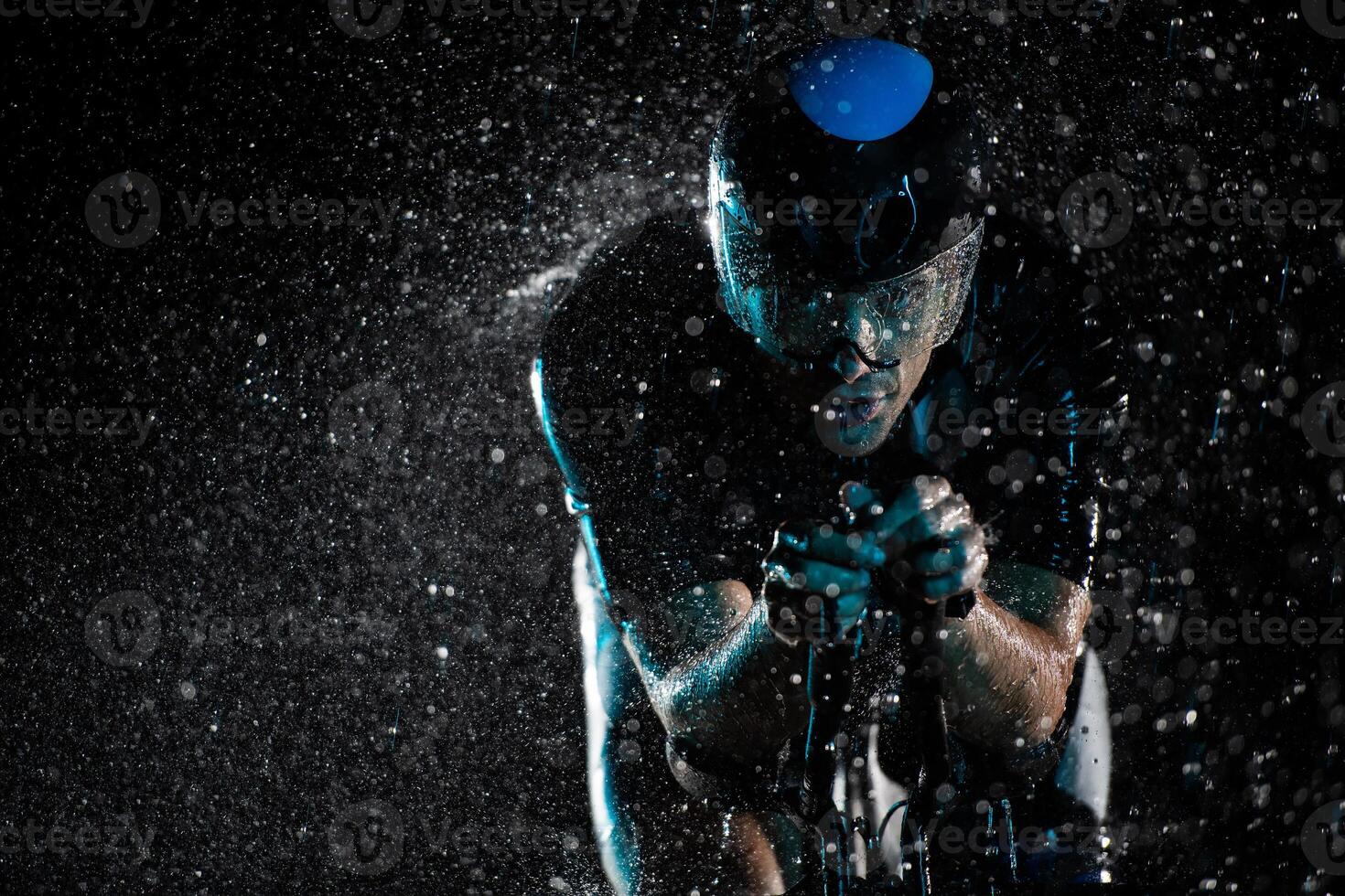 A triathlete braving the rain as he cycles through the night, preparing himself for the upcoming marathon. The blurred raindrops in the foreground and the dark, moody atmosphere in the background add photo