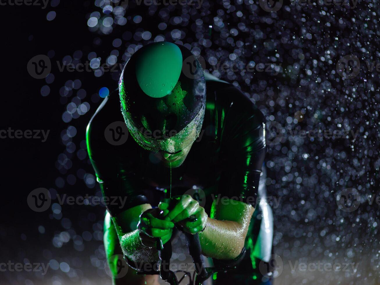 A triathlete braving the rain as he cycles through the night, preparing himself for the upcoming marathon. The blurred raindrops in the foreground and the dark, moody atmosphere in the background add photo