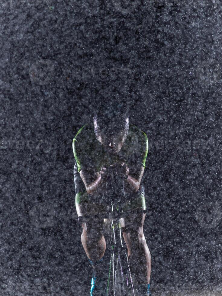 A triathlete braving the rain as he cycles through the night, preparing himself for the upcoming marathon. The blurred raindrops in the foreground and the dark, moody atmosphere in the background add photo