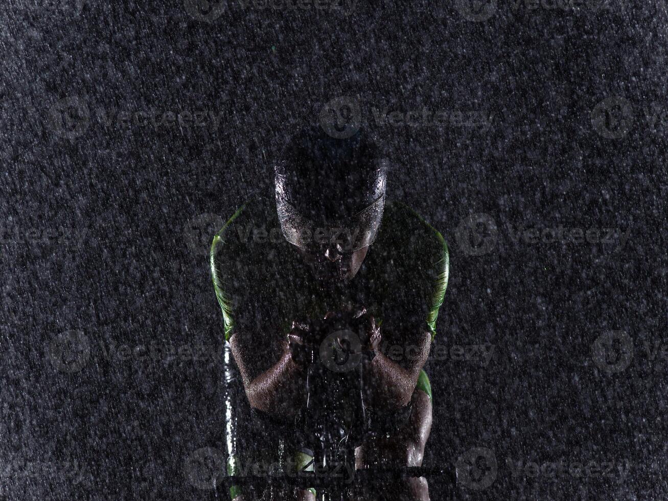 A triathlete braving the rain as he cycles through the night, preparing himself for the upcoming marathon. The blurred raindrops in the foreground and the dark, moody atmosphere in the background add photo