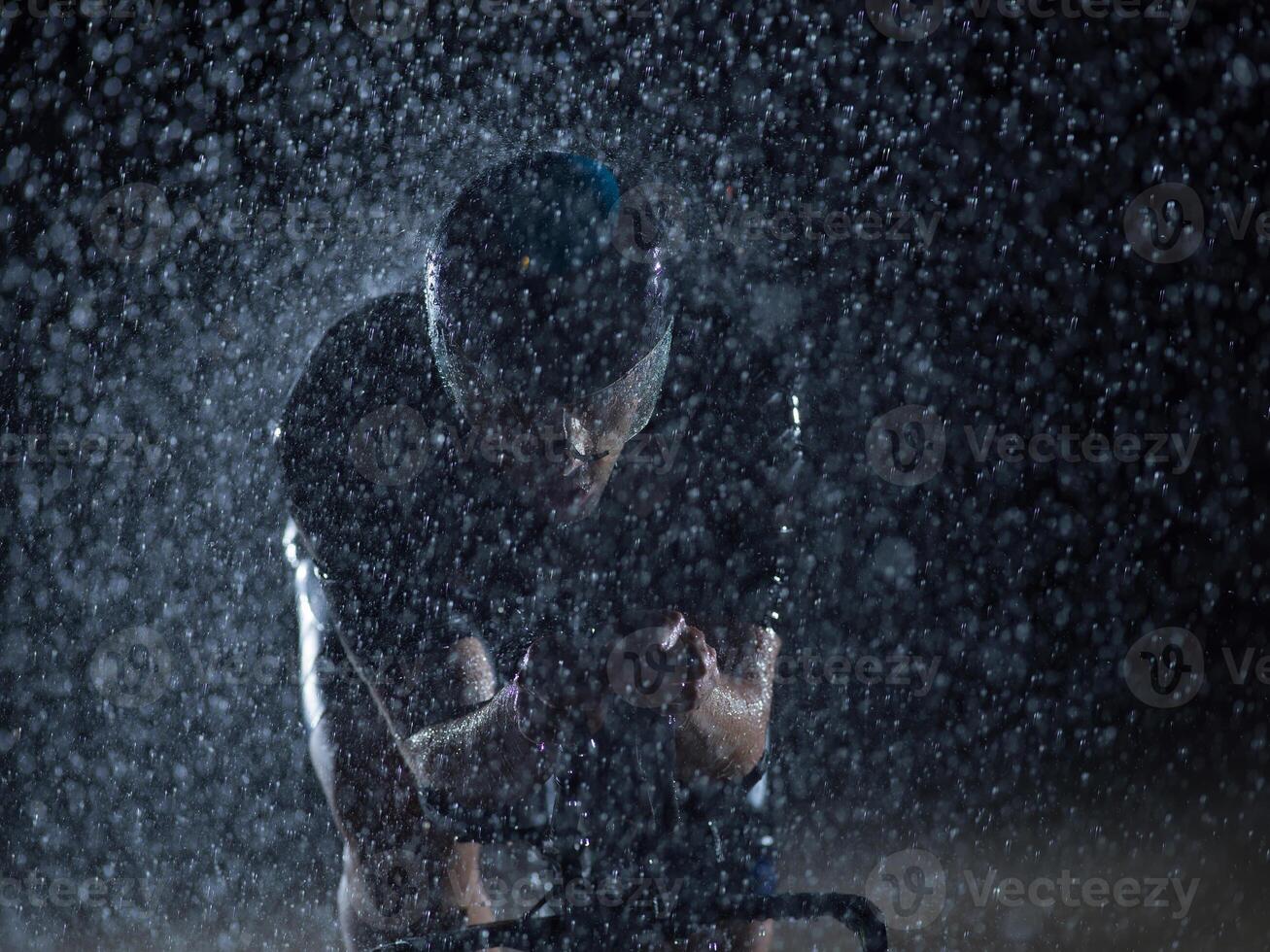 A triathlete braving the rain as he cycles through the night, preparing himself for the upcoming marathon. The blurred raindrops in the foreground and the dark, moody atmosphere in the background add photo