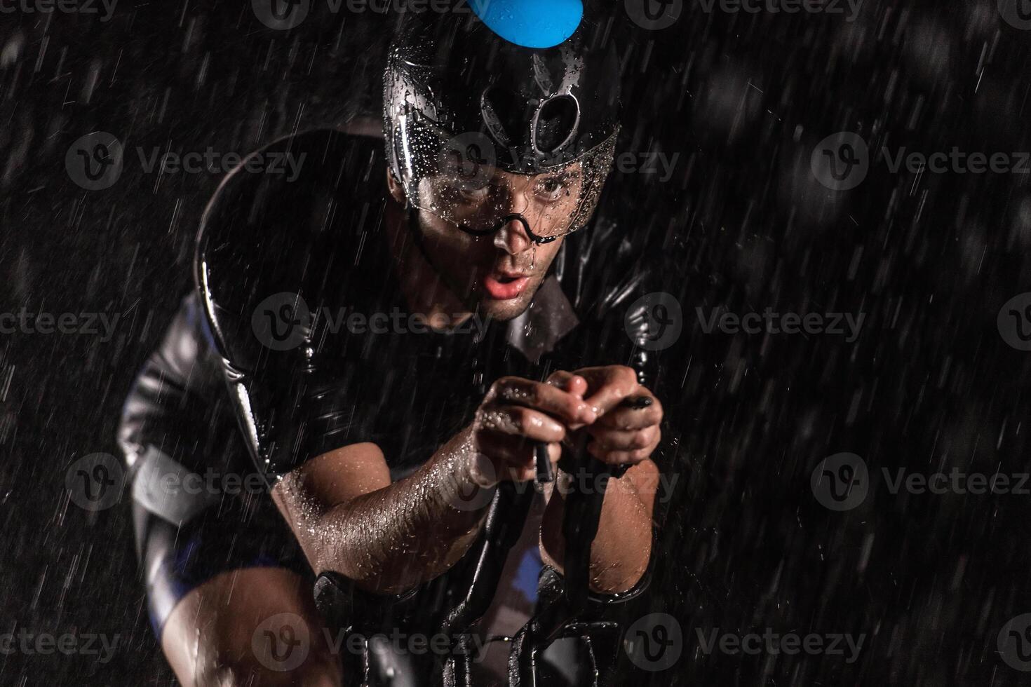 A triathlete braving the rain as he cycles through the night, preparing himself for the upcoming marathon. The blurred raindrops in the foreground and the dark, moody atmosphere in the background add photo