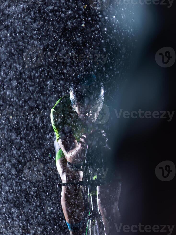 A triathlete braving the rain as he cycles through the night, preparing himself for the upcoming marathon. The blurred raindrops in the foreground and the dark, moody atmosphere in the background add photo