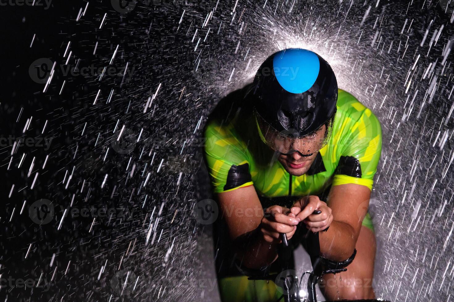 A triathlete braving the rain as he cycles through the night, preparing himself for the upcoming marathon. The blurred raindrops in the foreground and the dark, moody atmosphere in the background add photo