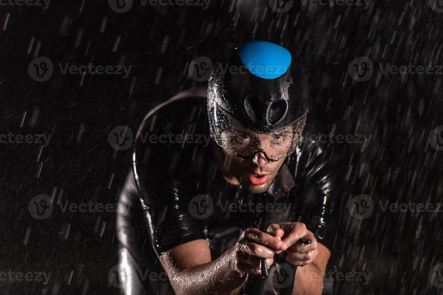 A triathlete braving the rain as he cycles through the night, preparing himself for the upcoming marathon. The blurred raindrops in the foreground and the dark, moody atmosphere in the background add photo