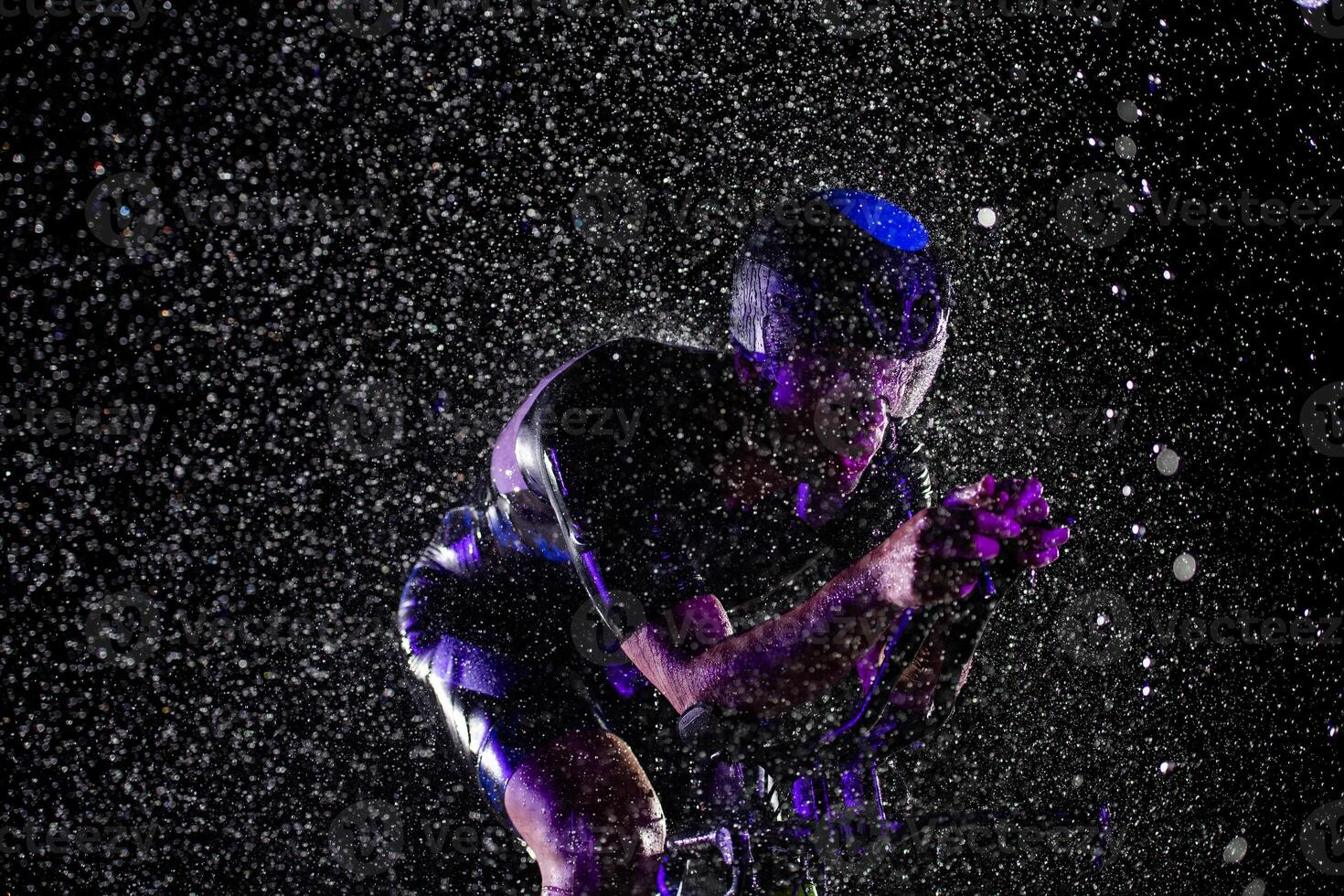 A triathlete braving the rain as he cycles through the night, preparing himself for the upcoming marathon. The blurred raindrops in the foreground and the dark, moody atmosphere in the background add photo