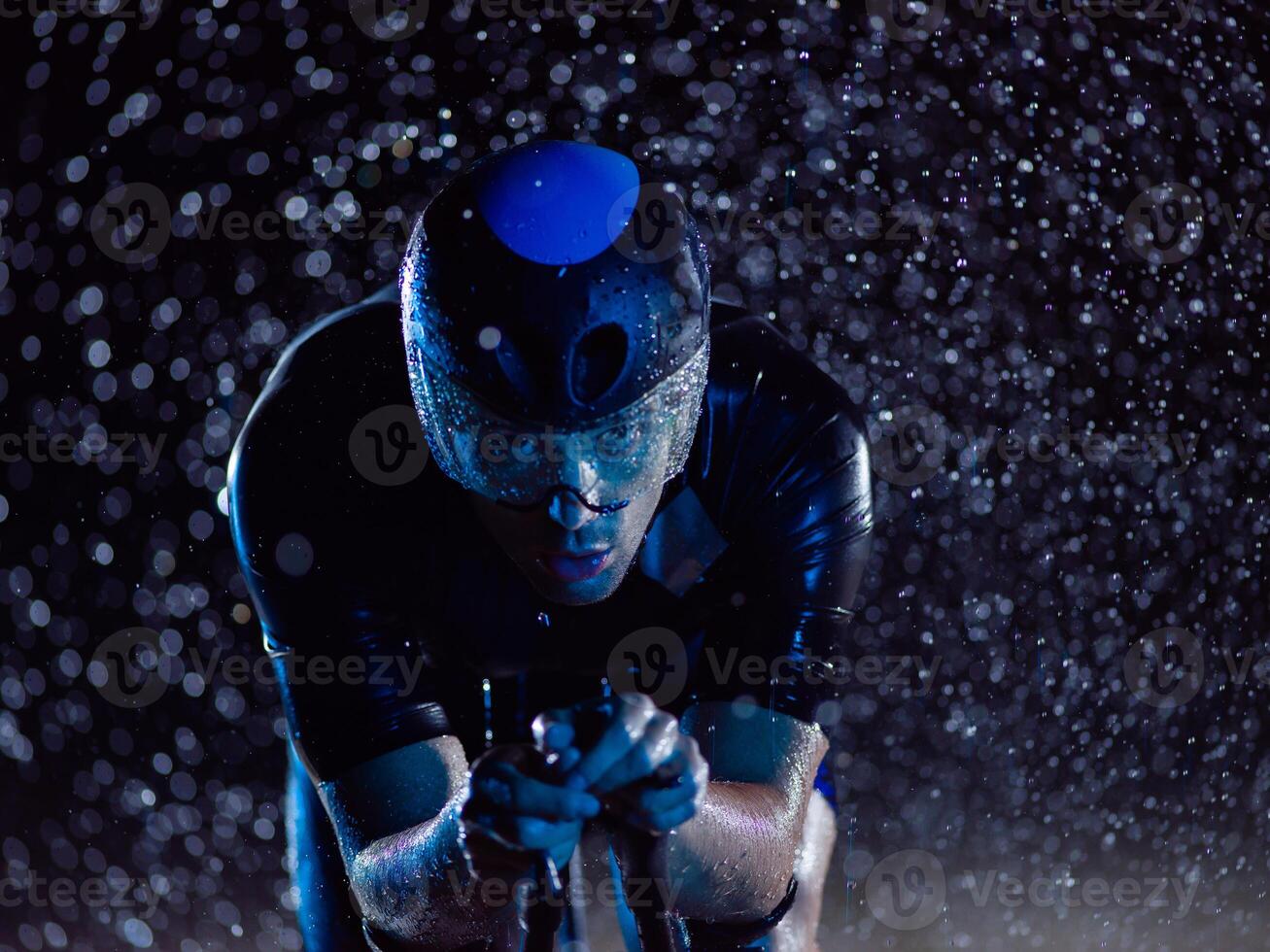 A triathlete braving the rain as he cycles through the night, preparing himself for the upcoming marathon. The blurred raindrops in the foreground and the dark, moody atmosphere in the background add photo