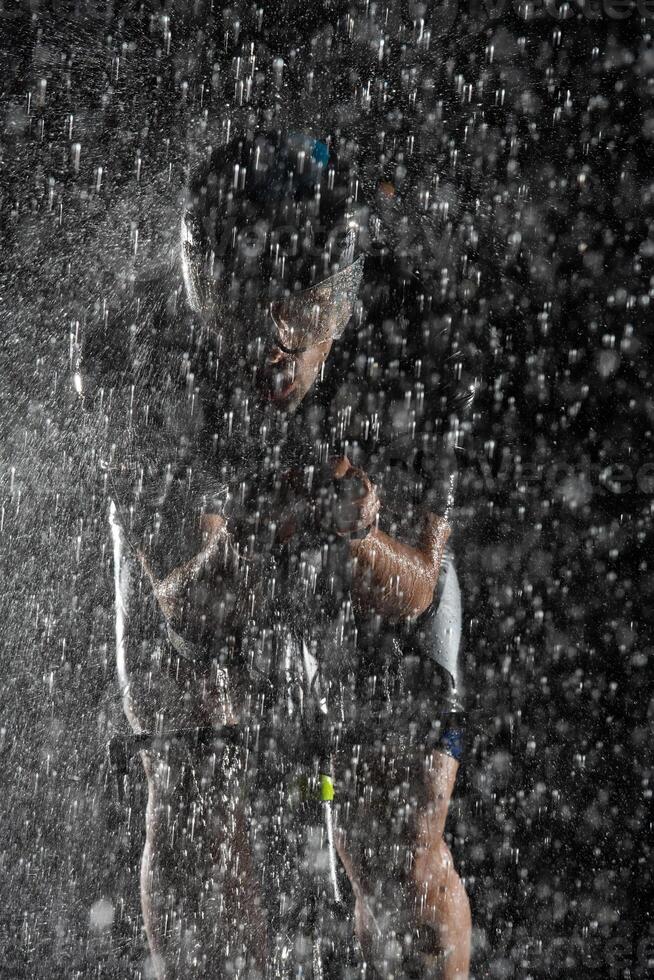 A triathlete braving the rain as he cycles through the night, preparing himself for the upcoming marathon. The blurred raindrops in the foreground and the dark, moody atmosphere in the background add photo