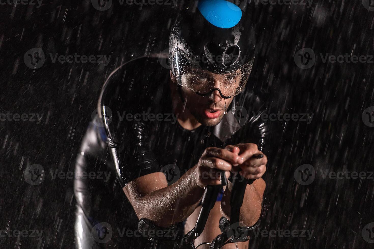 A triathlete braving the rain as he cycles through the night, preparing himself for the upcoming marathon. The blurred raindrops in the foreground and the dark, moody atmosphere in the background add photo