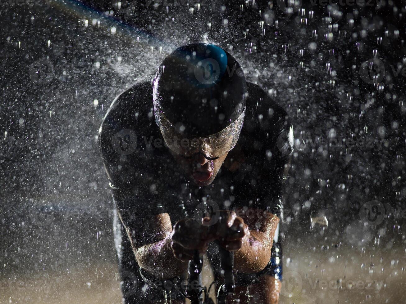 A triathlete braving the rain as he cycles through the night, preparing himself for the upcoming marathon. The blurred raindrops in the foreground and the dark, moody atmosphere in the background add photo