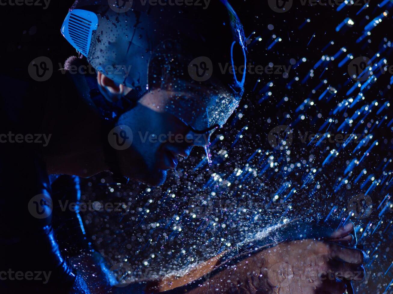 A triathlete braving the rain as he cycles through the night, preparing himself for the upcoming marathon. The blurred raindrops in the foreground and the dark, moody atmosphere in the background add photo