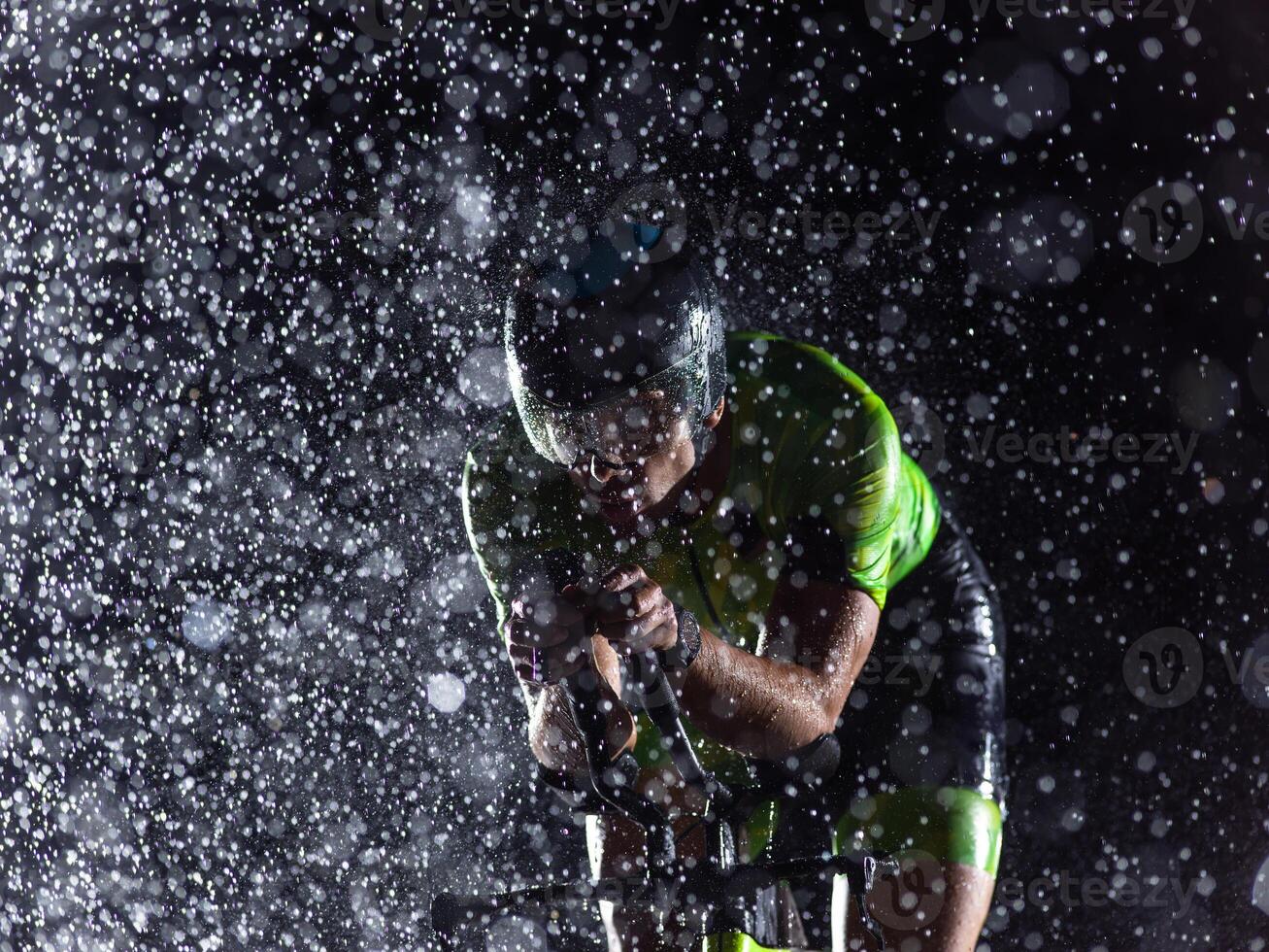 A triathlete braving the rain as he cycles through the night, preparing himself for the upcoming marathon. The blurred raindrops in the foreground and the dark, moody atmosphere in the background add photo