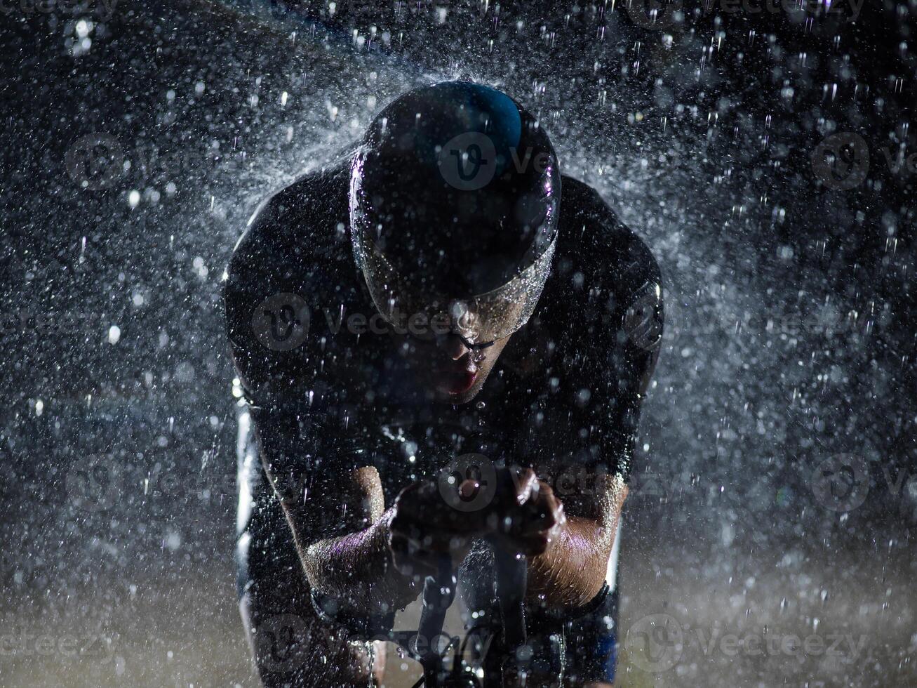 A triathlete braving the rain as he cycles through the night, preparing himself for the upcoming marathon. The blurred raindrops in the foreground and the dark, moody atmosphere in the background add photo