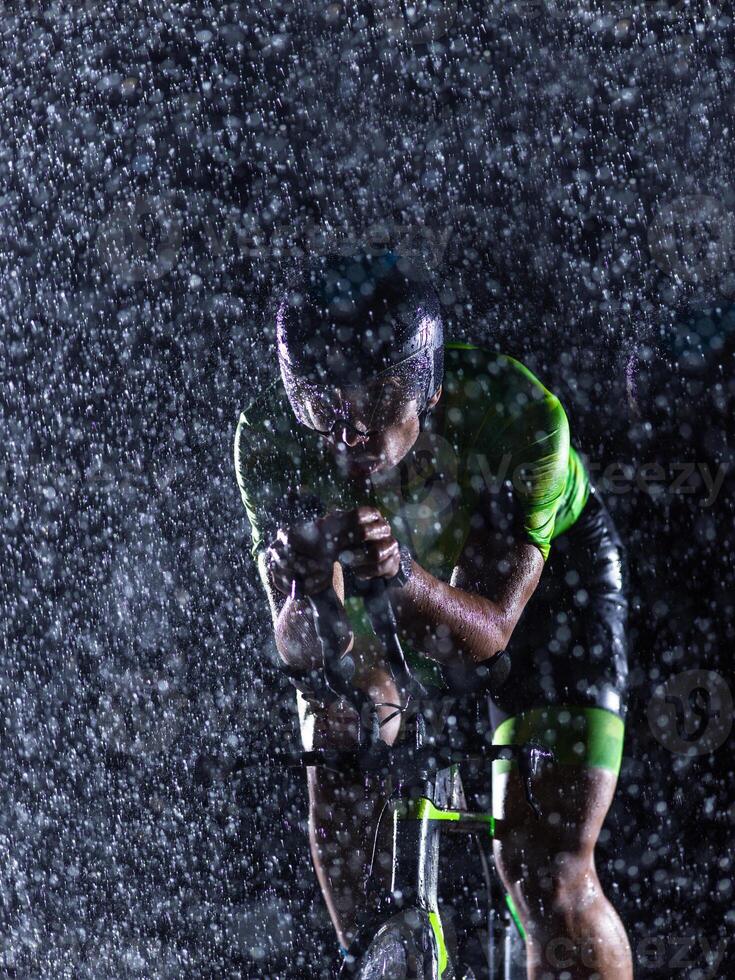 A triathlete braving the rain as he cycles through the night, preparing himself for the upcoming marathon. The blurred raindrops in the foreground and the dark, moody atmosphere in the background add photo