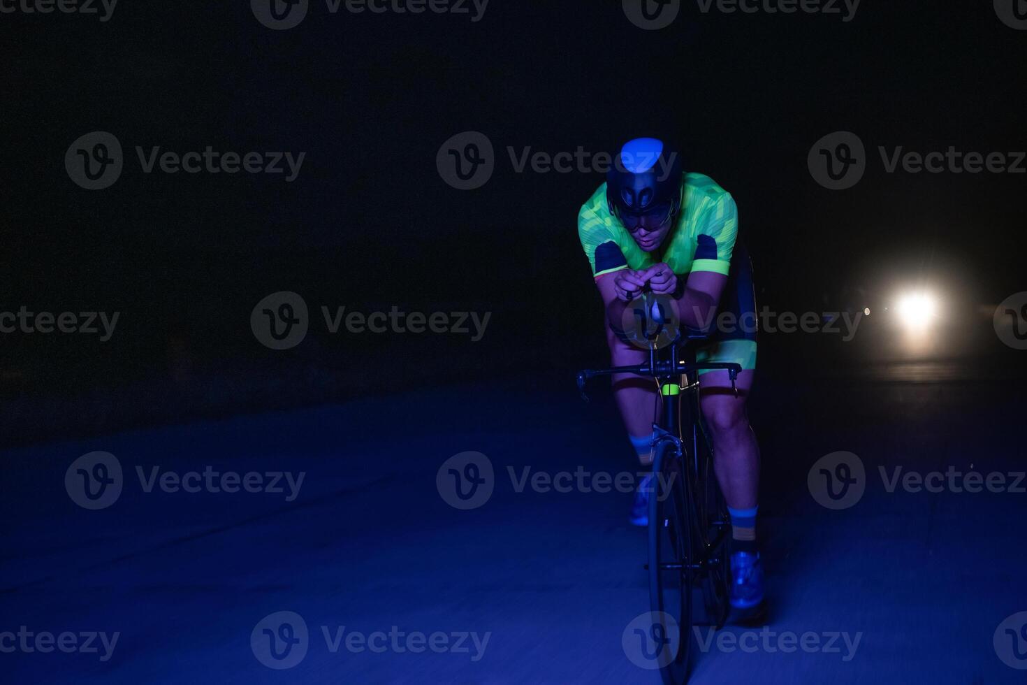 A triathlete rides his bike in the darkness of night, pushing himself to prepare for a marathon. The contrast between the darkness and the light of his bike creates a sense of drama and highlights the photo