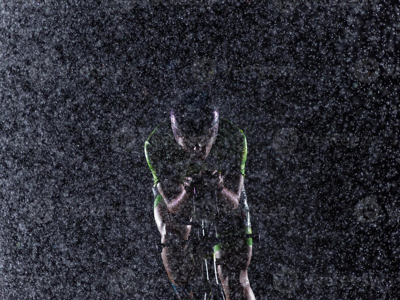 A triathlete braving the rain as he cycles through the night, preparing himself for the upcoming marathon. The blurred raindrops in the foreground and the dark, moody atmosphere in the background add photo