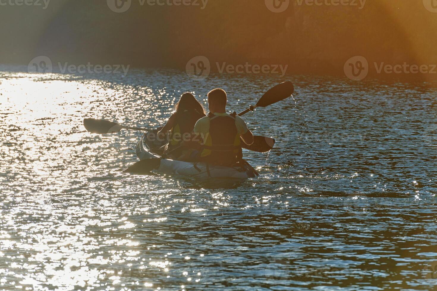 A young couple enjoying an idyllic kayak ride in the middle of a beautiful river surrounded by forest greenery in sunset time photo