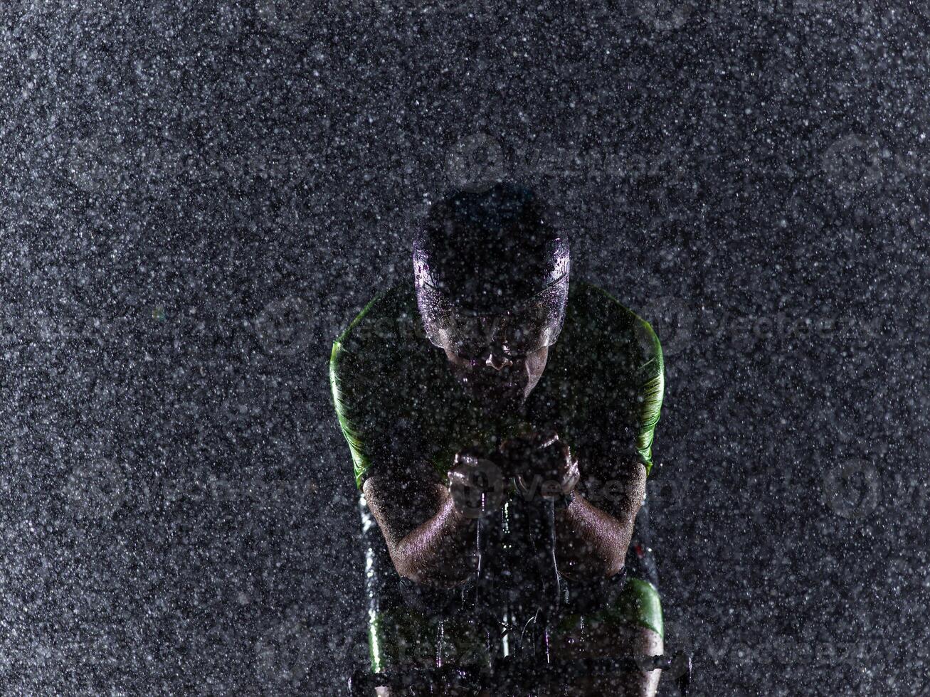 A triathlete braving the rain as he cycles through the night, preparing himself for the upcoming marathon. The blurred raindrops in the foreground and the dark, moody atmosphere in the background add photo