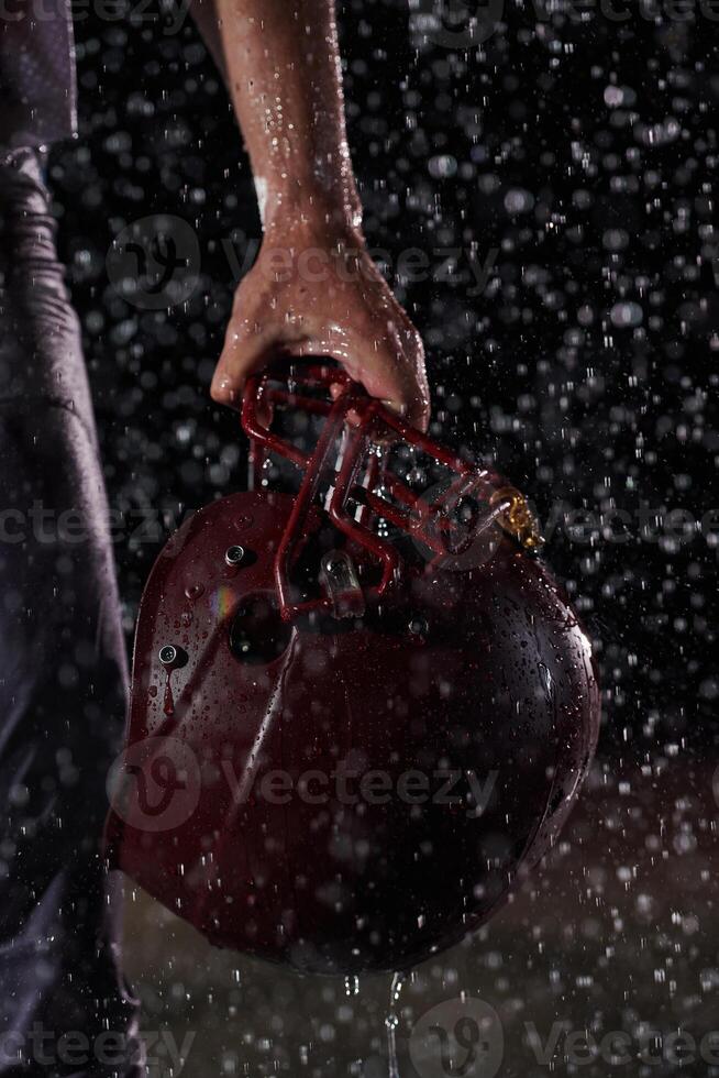 Close up of American Football Athlete Warrior Standing on a Field focus on his Helmet and Ready to Play. Player Preparing to Run, Attack and Score Touchdown. Rainy Night with Dramatic lens flare photo